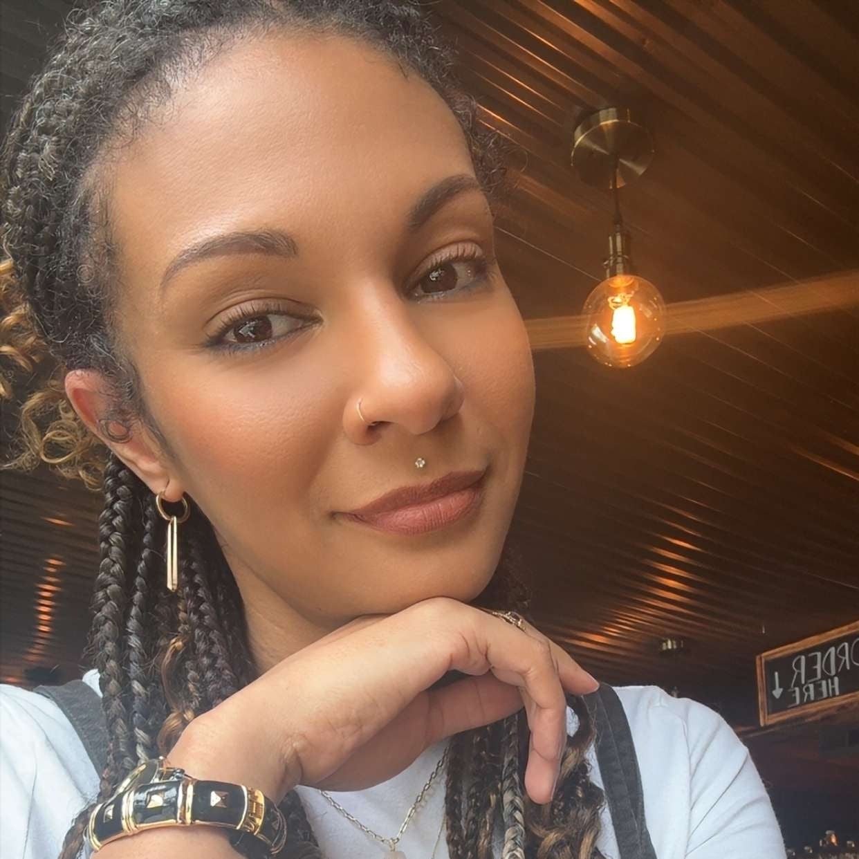 A close-up selfie of a woman with braided and curled hair featuring blonde and burgundy highlights, wearing a white t-shirt and dark gray apron inside what appears to be a salon or cafe.