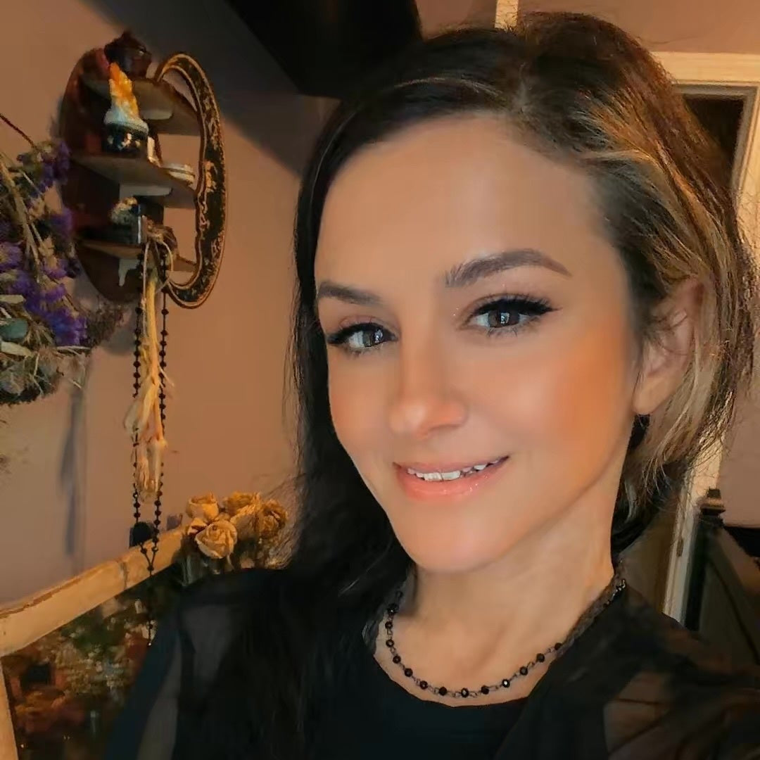 A woman with dark hair wearing a black sheer long-sleeve top and a black beaded necklace, posing for a selfie in an indoor setting with warm lighting.