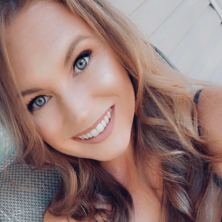 A smiling woman with long wavy light brown hair and blue eyes, wearing a black top, taking a selfie while leaning on a textured chair.